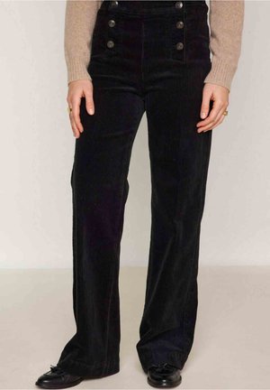 Femme portant un pantalon large en velours côtelé noir taille haute avec des boutons et des chaussures noires, debout devant un fond uni.