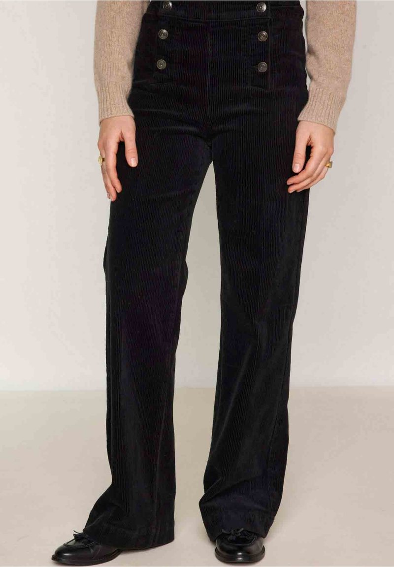 Femme portant un pantalon large en velours côtelé noir taille haute avec des boutons et des chaussures noires, debout devant un fond uni.