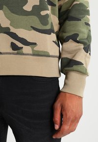 Felpa con motivo camouflage in beige, verde e nero. Polsini e vita a costine. Jeans neri con una texture liscia. Mano appoggiata sulla vita.