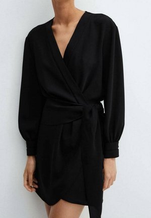 Kobieta ubrana w czarną sukienkę kopertową z długim rękawem, głębokim dekoltem w serek i przewiązana w talii, stojąca na jasnym, jednolitym tle.