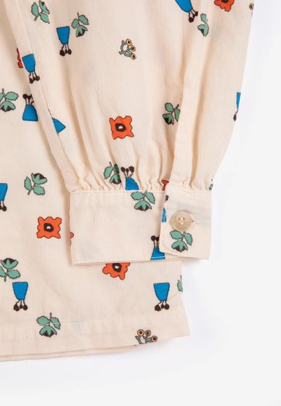 Manche en tissu beige avec imprimé floral coloré et bottes bleues, poignet froncé et fermeture par un seul bouton beige sur fond blanc.