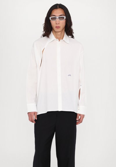 Chemise blanche à manches longues avec un col boutonné, dotée d'une fente sur les côtés et d'un petit logo bleu sur la poitrine. Portée avec un pantalon noir.