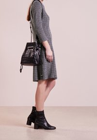 Donna con un vestito a fantasia fino al ginocchio, stivali neri alla caviglia e una borsa a tracolla in pelle nera con texture, in piedi su un pavimento semplice.