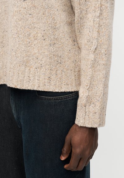 Μανίκι και μανσέτα μπεζ πουδρωμένου πλεκτού πουλόβερ στο μπράτσο ενός ατόμου που ακουμπάει δίπλα σε σκούρα μπλε τζιν.