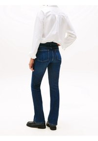 Personne portant une chemise blanche à manches longues, un jean bleu foncé ajusté avec des poches arrière, et des chaussures noires, debout avec une main sur la hanche, tournée vers l’arrière.