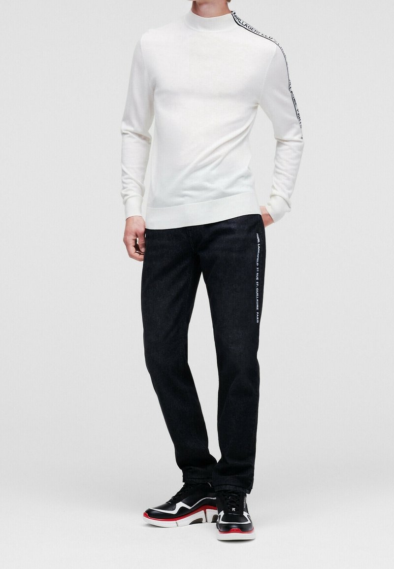 Homme portant un haut blanc à manches longues avec un texte sur l'épaule et un pantalon noir avec un texte sur la cuisse, associé à des baskets noir et blanc avec des accents rouges.