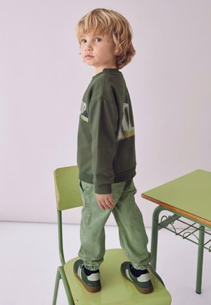 Felpa verde con una grafica colorata, abbinata a pantaloni verdi chiari e sneakers. Il bambino è in piedi su una sedia verde contro uno sfondo chiaro.