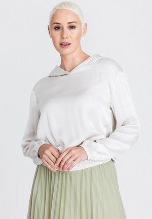 Frau mit kurzen blonden Haaren, die einen weißen Langarm-Hoodie und einen hellgrünen plissierten Rock trägt, steht vor einem einfarbigen Hintergrund.