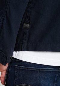 Mörkblå jacka med strukturerad tyg, med en G-Star-etikett, i kombination med en vit skjorta och mörka denimjeans med synlig sömnad.