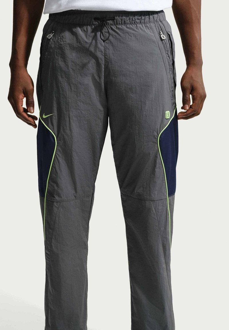 Persoon draagt grijze sportbroek met marineblauwe en neongroene details, handen ontspannen langs het lichaam, getoond van taille tot enkels tegen een effen achtergrond.