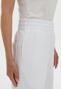 White cotton joggers with an elastic waistband, side pockets, and a soft texture, showcased next to a hand resting on the waistband.