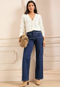 White knitted cardigan with a V-neck. Dark blue wide-leg jeans with braided pocket detailing. Beige suede handbag and heeled boots.