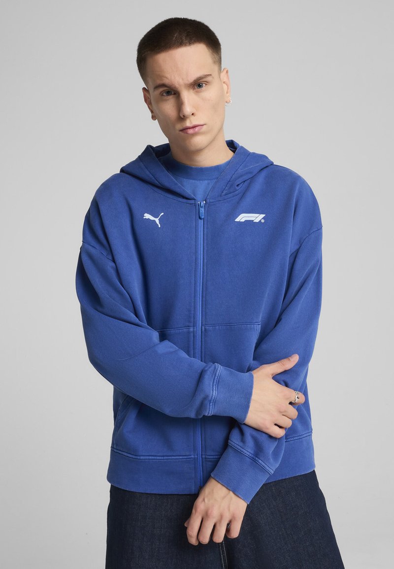 Plava zip-up dukserica od mekog materijala, s kapuljačom, kengur džepom, Puma logotipom i F1 brendiranim detaljima na prsima.