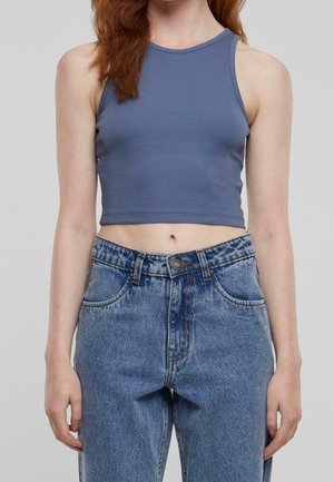 Femme portant un crop top bleu sans manches associé à un jean bleu taille haute et coupe ample sur un fond uni.