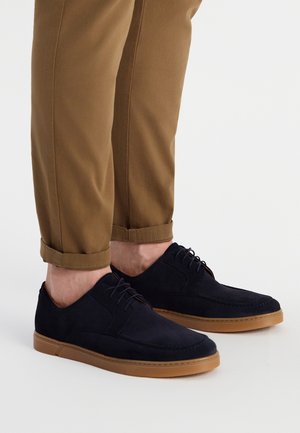 Zapatos de ante azul marino con un perfil bajo y suela de goma marrón. Presentan cordones y detalles de costura. Usados con pantalones beige arremangados.