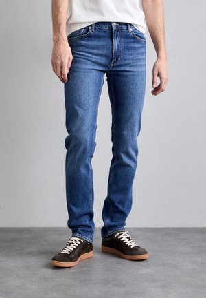 Denim jeans in a medium blue wash, slim fit, with five-pocket design, stitched detailing, and a smooth texture. Paired with dark suede sneakers.