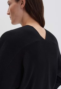 Frau mit braunem Haar, die einen schwarzen Pullover mit einem V-förmigen Rückenausschnitt trägt, von hinten und leicht von der Seite gesehen.