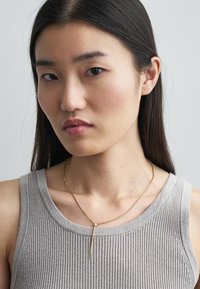 Χρυσή αλυσίδα κολιέ με λεπτό, κατακόρυφο μενταγιόν; το μοντέλο φοράει ένα ανοιχτό γκρι ριμπ αμάνικο μπλουζάκι. Απλός σχεδιασμός, γυαλιστερή επιφάνεια.