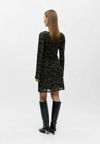 Moteris su ilgais tiesiais plaukais, vilkinti žalią permatomą suknelę su juodais gėlėtais raštais ir juodomis aukštakulnėmis botais iki kelių, atsisukusi atgal.