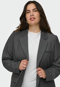 Šedý texturovaný blazer s klopovým límečkem a předními kapsami, nošený přes bílý žebrovaný top. Má volný střih a uvolněné rukávy.