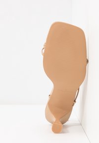 Sandale à talons hauts couleur nude avec une finition lisse et brillante, des lanières fines, un bout arrondi et une semelle en caoutchouc texturée.