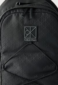 Μαύρο σακίδιο πλάτης Calvin Klein με διακριτικό επαναλαμβανόμενο μοτίβο λογότυπου, ελαστικό κορδόνι μπροστά και ορθογώνια ετικέτα με το λογότυπο στην πάνω πλευρά.