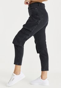 Pantalones cargo negros ajustados con múltiples bolsillos laterales, hechos de denim, con una cinturilla de altura media y dobladillos enrollados, combinados con zapatillas blancas.