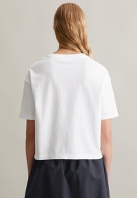 T-shirt blanc court en tissu côtelé. Manches courtes et col rond. Porté avec un vêtement sombre en couches.