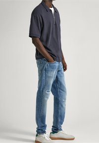 Marinblå zip-up polo, mjukt tyg; ljusblå slitna jeans; vita och gröna sneakers med gummisulor; avslappnad passform med uppvikt ärmar.