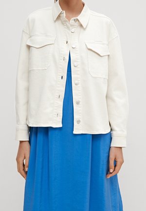 Femme portant une veste blanche à boutons ouverte sur une robe bleu vif, visible du cou au milieu des cuisses sur un fond uni.