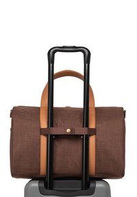 Hnědý duffel bag s koženými akcenty, připevněný k rukojeti černého kufru. Má texturovanou látku a detaily ze zlaceného kovu.