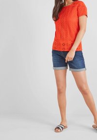 Punainen virkatut lyhythihainen paita, jossa on kukkaleikkauksia ja aallotettu helma, yhdistettynä käännettyihin farkkushortseihin ja liukuviin sandaaleihin.