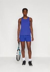 Top desportivo azul e calções, com textura suave. Usado com sapatilhas desportivas pretas e segurando uma raqueta de ténis. Meias brancas até ao tornozelo.