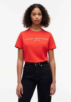 Kobieta z kręconymi włosami, ubrana w czerwoną koszulkę Tommy Hilfiger i ciemne dżinsy, stojąca na tle jednolitego białego tła.