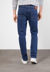 Jean en denim bleu avec une coupe droite, comportant deux poches arrière et une étiquette en cuir marron à la taille. Porté avec des baskets blanches.