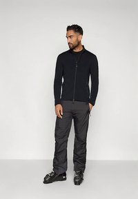 Άνδρας που φορά μαύρο μπουφάν με φερμουάρ, μαύρο παντελόνι και μαύρες μπότες σκι, στέκεται με το ένα χέρι στην τσέπη, κοιτώντας στο πλάι.