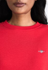 Red sweatshirt with ribbed crew neckline, featuring a small embroidered logo on the chest. Fabric appears soft with a smooth texture.
