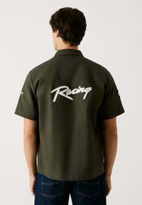 Camisa de manga corta verde oliva con collar abotonado, que presenta "Racing" bordado en blanco en la parte posterior y un parche de logo en la manga.