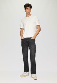 Witte katoenen t-shirt, donkere spijkerbroek en lichte sneakers. Model staat met handen in de zakken en toont een casual look met een neutrale achtergrond.
