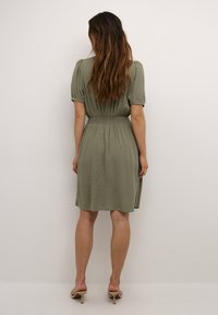 Olijfgroene jurk met pofmouwen, smock taille en subtiele textuur, tot net boven de knie vallend. Draagt met lichtgekleurde hoge hakken.
