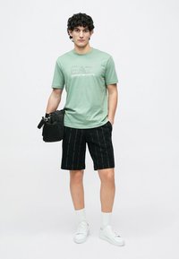 Vaaleanvihreä puuvillainen t-paita, jossa on "EA7 Emporio Armani" -logo, yhdistettynä mustiin shortseihin, joissa on pystysuoria tekstikuoseja, ja valkoisiin lenkkareihin.