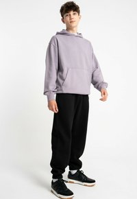 Jasnofioletowa bluza z kapturem z kieszenią kangurzą, w połączeniu z czarnymi dresami. Model nosi czarne sneakersy z białą podeszwą.