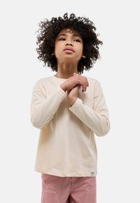 T-shirt en coton beige à manches longues avec un col rond, coupe décontractée et poignets côtelés. Porté avec un pantalon rayé rouge et blanc.
