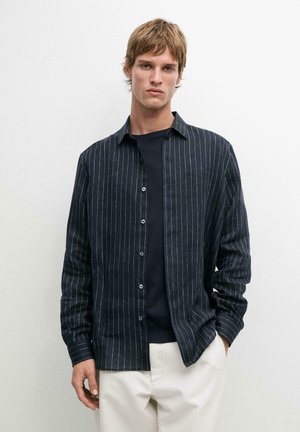 Junger Mann, der ein schwarzes Nadelstreifenhemd über einem schwarzen T-Shirt und weißen Hosen trägt, vor einem schlichten weißen Hintergrund stehend.