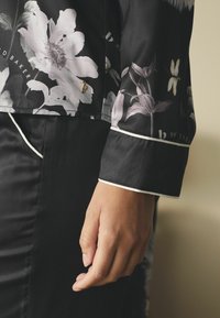 Article de îmbrăcăminte din satin negru cu imprimeu floral în nuanțe de alb și gri. Prezintă finisaje albe la manșete și un accent auriu la tiv.
