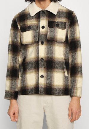 Person trägt eine braun-schwarz-creme karierte Jacke mit Knöpfen, zwei Frontklapptaschen und einem cremefarbenen Sherpa-Kragen.