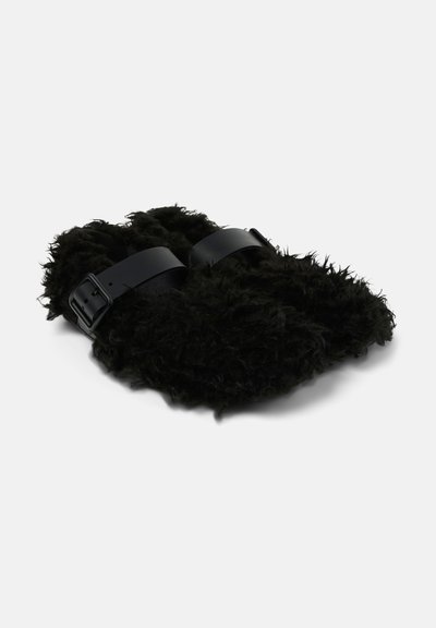 Pantofole nere in peluche con due cinturini neri regolabili, inclinate per mostrare la parte anteriore e laterale su uno sfondo bianco.