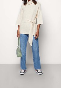 Personne portant un pull à col roulé crème avec ceinture à la taille, jean bleu, baskets noires et blanches, tenant un petit sac à main vert clair.