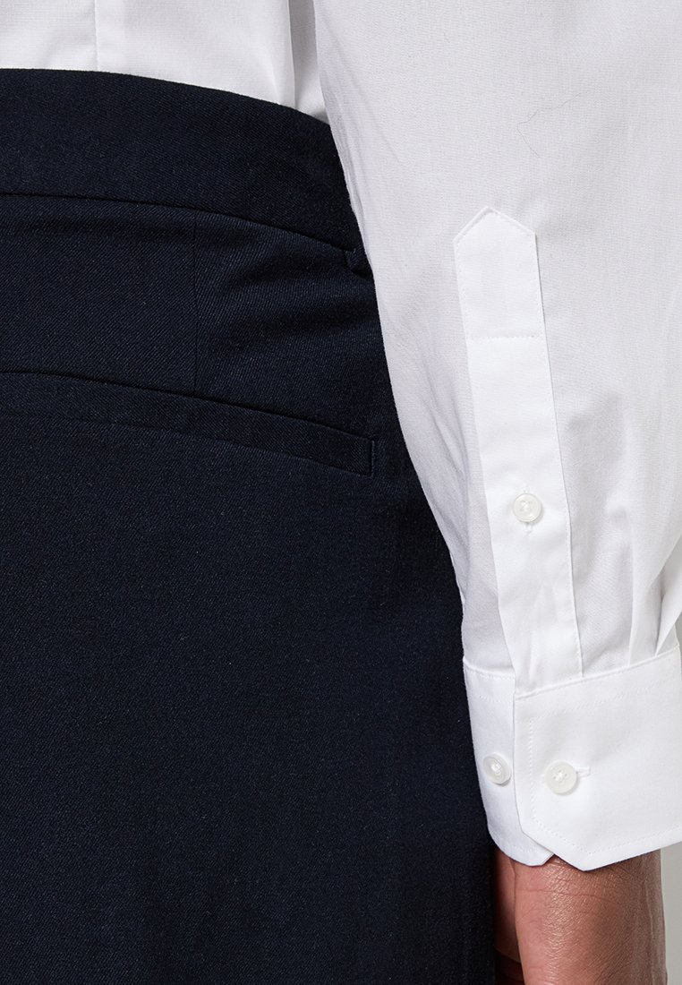 Manga de camisa blanca de cuello junto a pantalones azul marino; presenta cierres de botón, textura de tela suave y diseño ajustado.