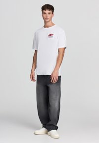 Biała bawełniana koszulka z luźnym krojem i krótkimi rękawami, z czerwoną grafiką na piersi; w zestawieniu z czarnymi dżinsami i białymi trampkami.